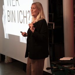 Susanne Maisch spricht vor Leinwand, auf der steht "Wer bin ich?"